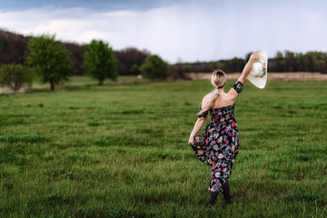 a beautiful girl in a dress with a hat in her hand walks alone in a green meadow. Young pretty blond woman in nature runs forward and enjoys freedom and relaxation