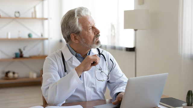 Serious senior doctor physician looking away thinking of future healthcare challenges at workplace with laptop. Thoughtful old adult male therapist feeling concerned about medicine problem solution.