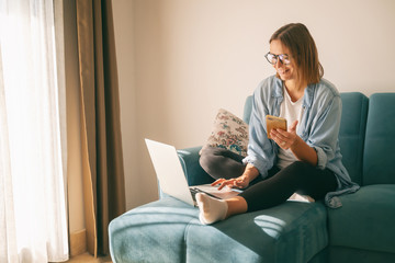 Young mature attractive woman sitting on a blue sofa in the living room at home with a smartphone in her hands in front of a laptop, online shopping