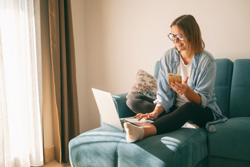 Young mature attractive woman sitting on a blue sofa in the living room at home with a smartphone in her hands in front of a laptop, online shopping - Powered by Adobe