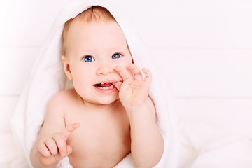 Cute baby of 8 months in a big white towel is dressed on his head. Holds fingers in the mouth. Clean little baby after bath.