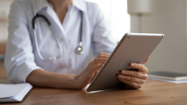 Female doctor physician wearing white coat holding modern digital tablet computer in hands using healthcare medical tech app working in software web program at workplace in hospital. Close up view.