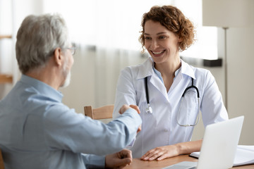Fototapeta premium Smiling young female doctor handshaking greeting healthy senior male patient in hospital. Happy practitioner shake hand of elder client at advice meeting. Old people medical health care trust concept.