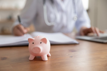 Female doctor working at desk with piggy bank box. Piggybank on table as concept of healthcare fees financial cost, money savings on health care insurance, medical care expenses concept. Close up view
