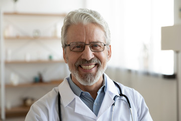 Smiling professional older man doctor wears white coat, glasses and stethoscope looking at camera. Happy bearded senior physician or therapist with dental smile posing for close up head shot portrait.