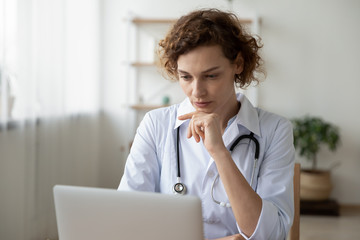 Concentrated young female physician working on computer looking at laptop screen thinking on...