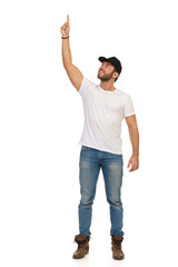 Young Man In Jeans, White T-shirt And Black Cap Is Standing With Arm Raised And Pointing Up