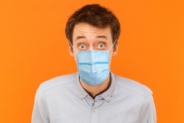 Closeup portrait of shocked young worker man with surgical medical mask standing and looking at camera with big eyes. indoor studio shot isolated on orange background.