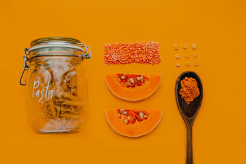 A glass jar of pasta, dry red lentils, slices of butternut squash pumpkin, sweetcorn and a wooden spoon of turmeric on a yellow background in a flat lay composition