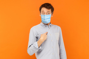 Who? me? Portrait of shocked young worker man with surgical medical mask standing pointing himself, asking and looking at camera with amazed face. indoor studio shot isolated on orange background.