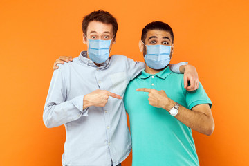 Portrait of two young worker men with surgical medical mask standing, hugging, showing and pointing each other and looking at camera with funny face. indoor studio shot isolated on orange background.