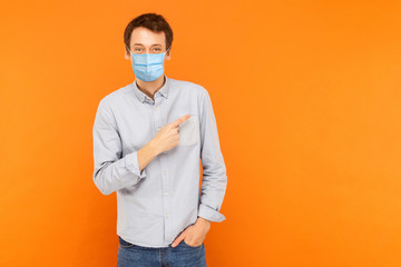 Portrait of happy young worker man with surgical medical mask standing, pointing and showing background empty copy space and smiling. indoor studio shot isolated on orange background.