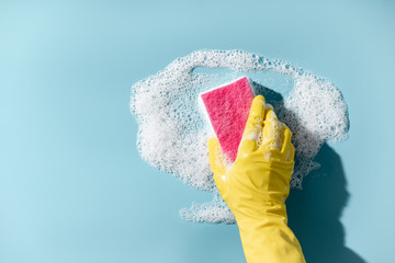 Hand in a yellow rubber glove holds a cleaning sponge and wipes a soapy foam on a blue background. Cleaning concept, cleaning service - obrazy, fototapety, plakaty