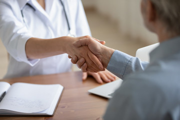 Female doctor physician and senior adult male patient shake hands with handshake greeting over hospital table. Elderly health care medical services trust, gratitude, agreement concept. Close up view