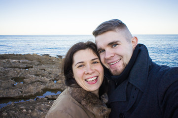 Obraz premium Portrait of young beautiful couple taking selfie photo in a smartphone with sea and dark cloudy sky on the background. Cold season and travel concept.