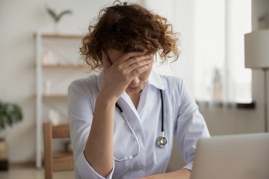 Exhausted Depressed Young Female Doctor Feels Mental Burnout At Work. Stressed Frustrated Physician Upset About Medical Failure, Healthcare Negligence, Having Headache Sits Alone At Desk In Hospital.