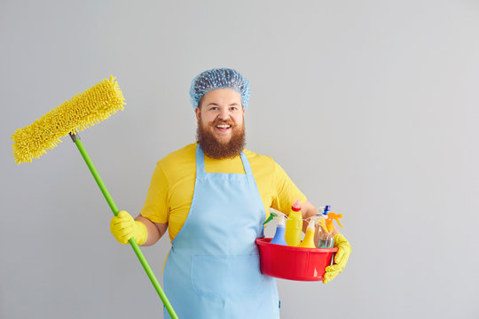 Funny Fat Man In An Apron And Yellow Cleaning Gloves Is Cleaning On Gray Background.