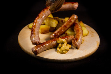 kitchen tongs resting an exquisite Italian sausage cooked on the grill with browned diced potatoes placed on a wooden cutting board with drops of mustard on a black background
