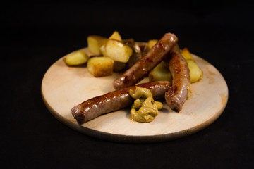 delicious Italian sausage cooked on the grill with browned diced potatoes placed on a wooden cutting board with drops of mustard with a black background 
