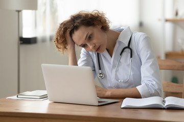 Stressed young female doctor looking at laptop reading bad news online worried of mistake at...
