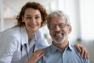 Happy young adult female doctor physician wearing white coat and stethoscope embracing smiling healthy senior adult male patient looking at camera. Elderly people medical health care concept. Portrait