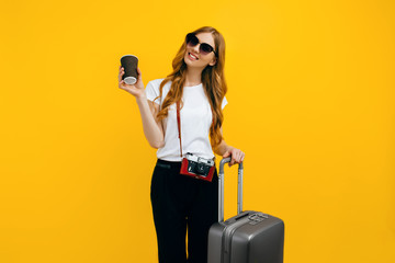 excited tourist woman with a retro camera and a suitcase, drinking coffee on a yellow background. Concept of going abroad, traveling