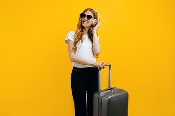 A young smiling tourist, with a suitcase listening to music on headphones on a colorful yellow background