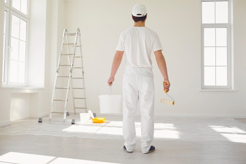 A male painter in a white uniform with a roller works in his hand in a white room - obrazy, fototapety, plakaty