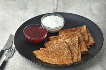 Thin pancakes on a black plate.  Cups with sour cream and jam next to pancakes.  Light background.  Close-up.