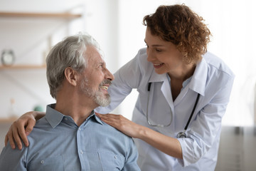 Kind smiling female doctor embracing encouraging happy senior male patient in hospital. Happy healthy older man and his physician enjoying talking at nursing home. Elderly medical health care concept.