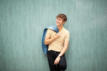 smiling young man posing by green wall with denim jacket over shoulder