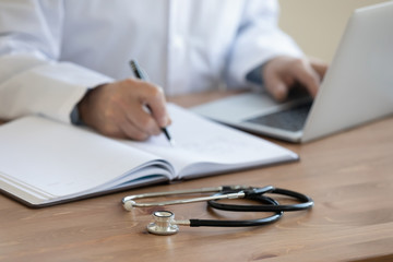 Fototapeta premium Senior doctor physician using laptop, writing prescription with stethoscope on desk. Old male professional therapist wearing white coat working in hospital. Healthcare, medicine concept. Close up view
