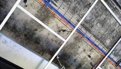 Cable tray with wires and metal duct show above t-bar grid in opened suspended (drop) ceiling and below concreate of upper floor at construction site of industrial building - obrazy, fototapety, plakaty