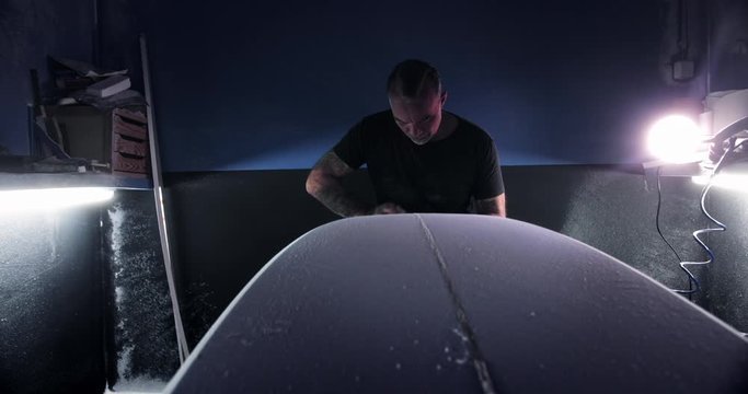 Man shaping surfboard with sanding block in workshop