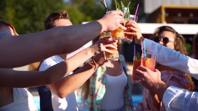 Group of friends having fun at poolside summer party taking colorful cocktails from table and clinking glasses smiling. People toast drinking fresh juice at luxury tropical villaon summer day.