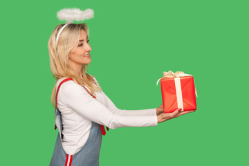 Happy birthday! Side view of angelic adult woman with halo above head giving gift box and smiling,...