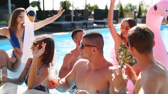 Friends having party with cocktails in holiday villa swimming pool. Happy young people in swimwear dancing, clubbing with inflatable flamingo, swan, mattress in luxury resort on sunny day. Slow motion