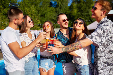 Group of friends having fun at poolside summer party clinking glasses with summer cocktails on sunny day near swimming pool. People toast drinking fresh juice at luxury villa on tropical vacation.