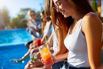 Pretty woman relax with fresh colorful cocktail glass sitting by swimming pool on sunny summer day with friends. People toast drinking beverages at luxury villa poolside party on tropical vacation.