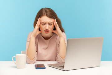 Frustrated tired young woman employee sitting at workplace with laptop and holding painful head,...