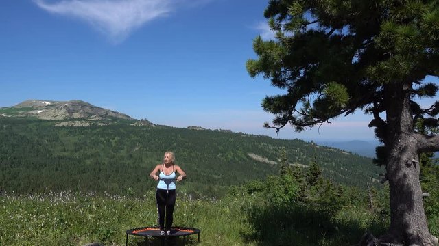 a young woman jumps on a trampoline against the background of a summer mountain valley at a summer resort. outdoor aerobics classes. outdoor fitness, Sports and healthy lifestyle. Pretty fit girl
