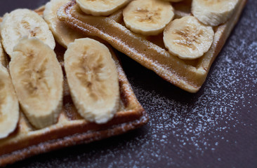 Belgian waffles with banana and sugar powder on dark background.