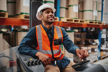 Happy forklift driver focused on carefully transporting stock from shelves around the floor of a large warehouse wearing a white helmet and vest © StratfordProductions