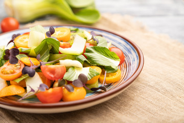 Vegetarian salad of pac choi cabbage, kiwi, tomatoes, kumquat, microgreen sprouts on gray wooden background. Side view, selective focus.