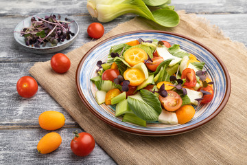 Vegetarian salad of pac choi cabbage, kiwi, tomatoes, kumquat, microgreen sprouts on gray wooden background. Side view, close up.