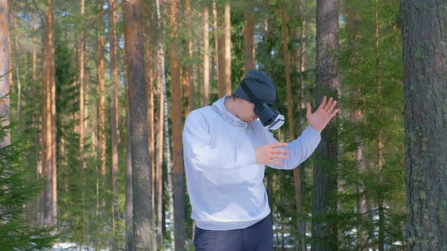 Portrait man in forest among pine trees in vr googles waving hands and turning sides. Playing games or watching 3d films on virtual reality headset in Augmented Reality. Challenge on street in spring.