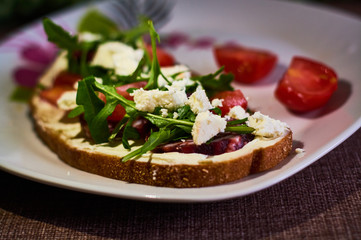 Large toast of rye bread with pieces of cheese, arugula, cherry tomatoes and sausage.
