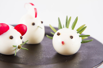 Eggs shaped like animals (with elements made of rosemary, radish, chive) on a plate, on a white background