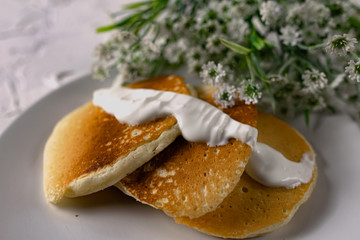 Crumpets and pancakes with sour cream on a white plate with white sauce on top. Next to a sprig of fresh light flowers and compote. Interesting textured background of white stone