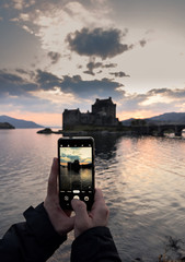 Cropped boy hands taking a smart phone photo to an old scottish castle surrounded by water in a sunset